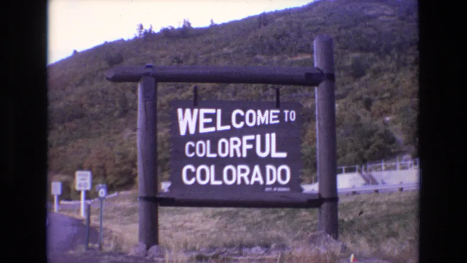 Final 1973 8mm frame holding on the Welcome to Colorful Colorado roadside sign, with traffic signs, guardrail, grass, and mountain foliage visible.