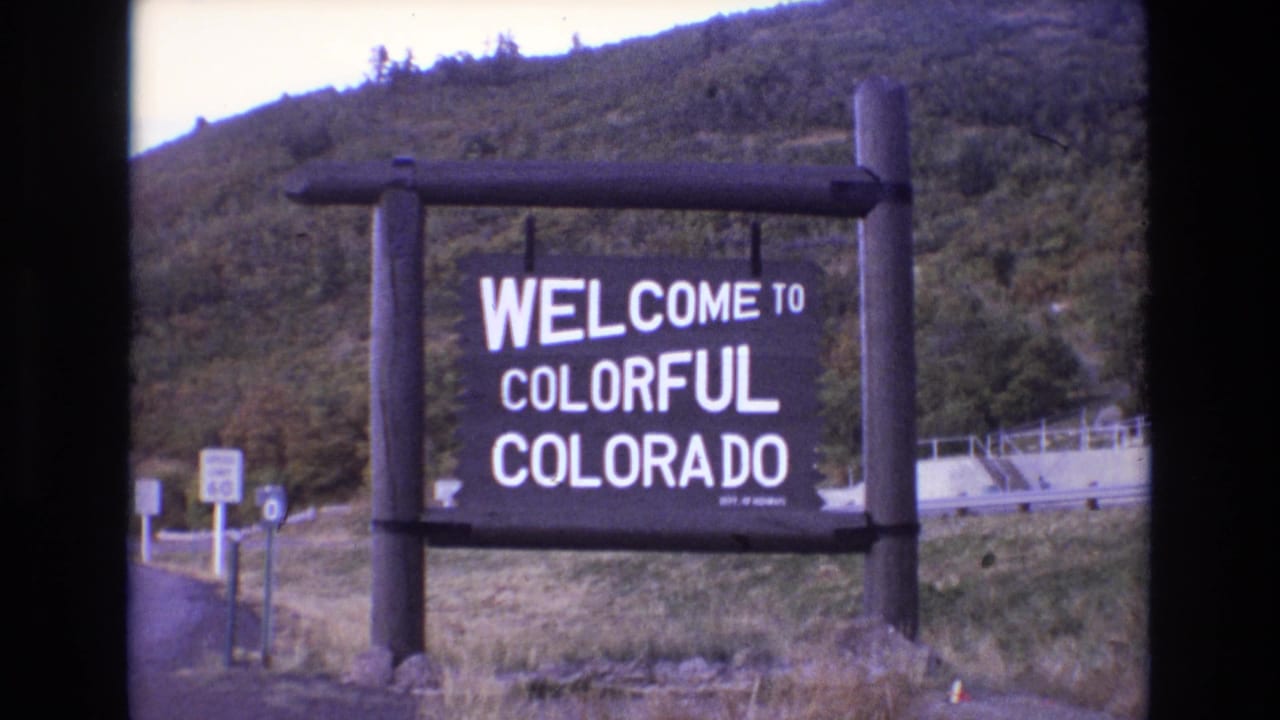 1973 8mm frame of a Welcome to Colorful Colorado sign on wooden posts beside a roadside shoulder with green mountain terrain behind it.