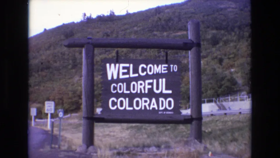 1973 8mm frame showing the Welcome to Colorful Colorado sign from a slightly shifted angle, with wooden supports, roadside signage, and a wooded hillside in the background.
