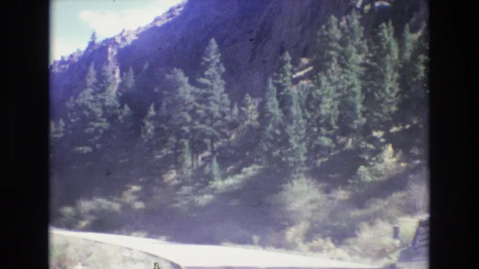 1973 8mm frame looking along a winding Colorado mountain road bordered by pine trees, brush, and shadowed slopes under daylight.