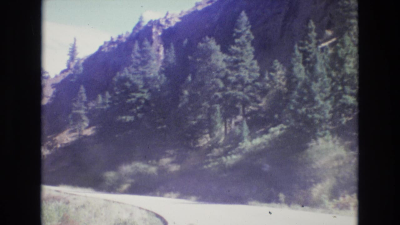 1973 Colorado 8mm frame showing a curving mountain road beneath pine trees and steep rocky terrain.