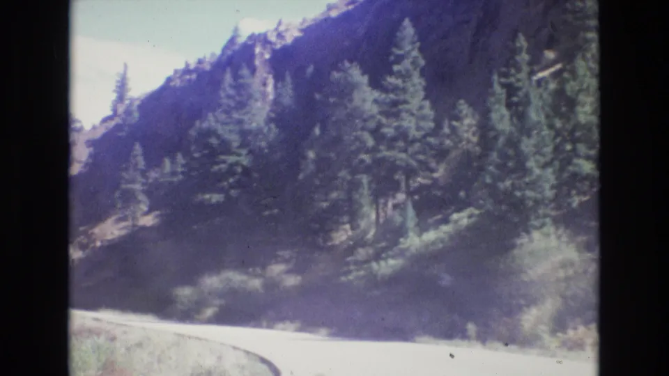 1973 Colorado 8mm frame showing a curving mountain road beneath pine trees and steep rocky terrain.