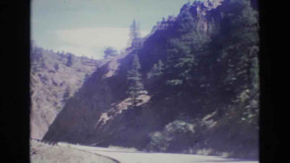 1973 Colorado home-movie frame of a road bend cutting through rocky mountain terrain with evergreens above it.
