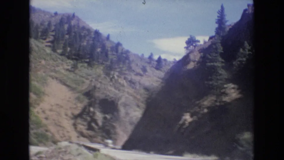 1973 Colorado 8mm view of a winding road at the base of a steep slope with scattered pines and bright sky.