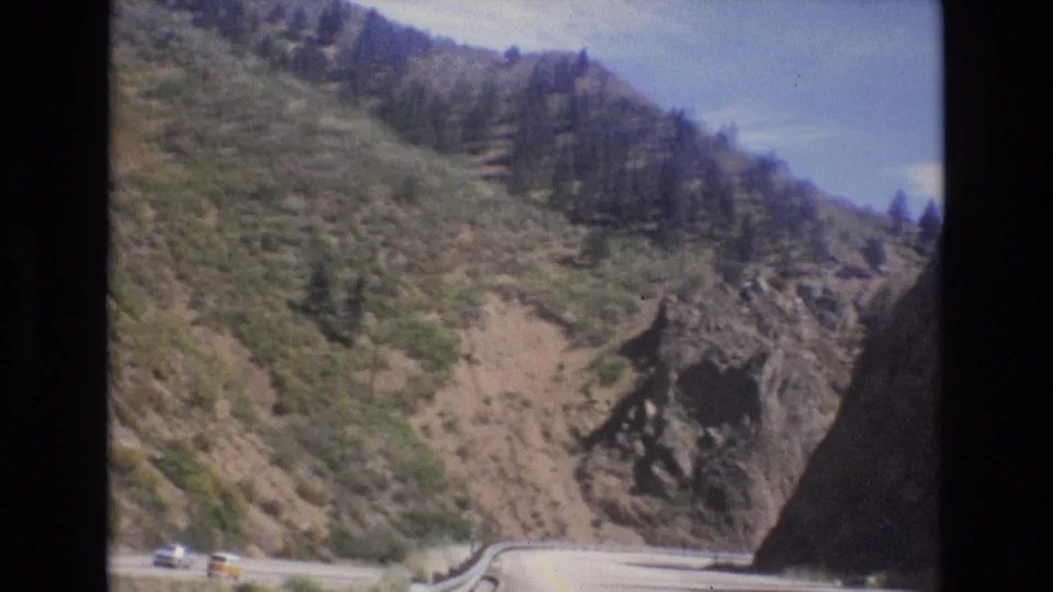 1973 Colorado home-movie frame showing small cars traveling along a winding road under a steep, partly forested mountain slope.