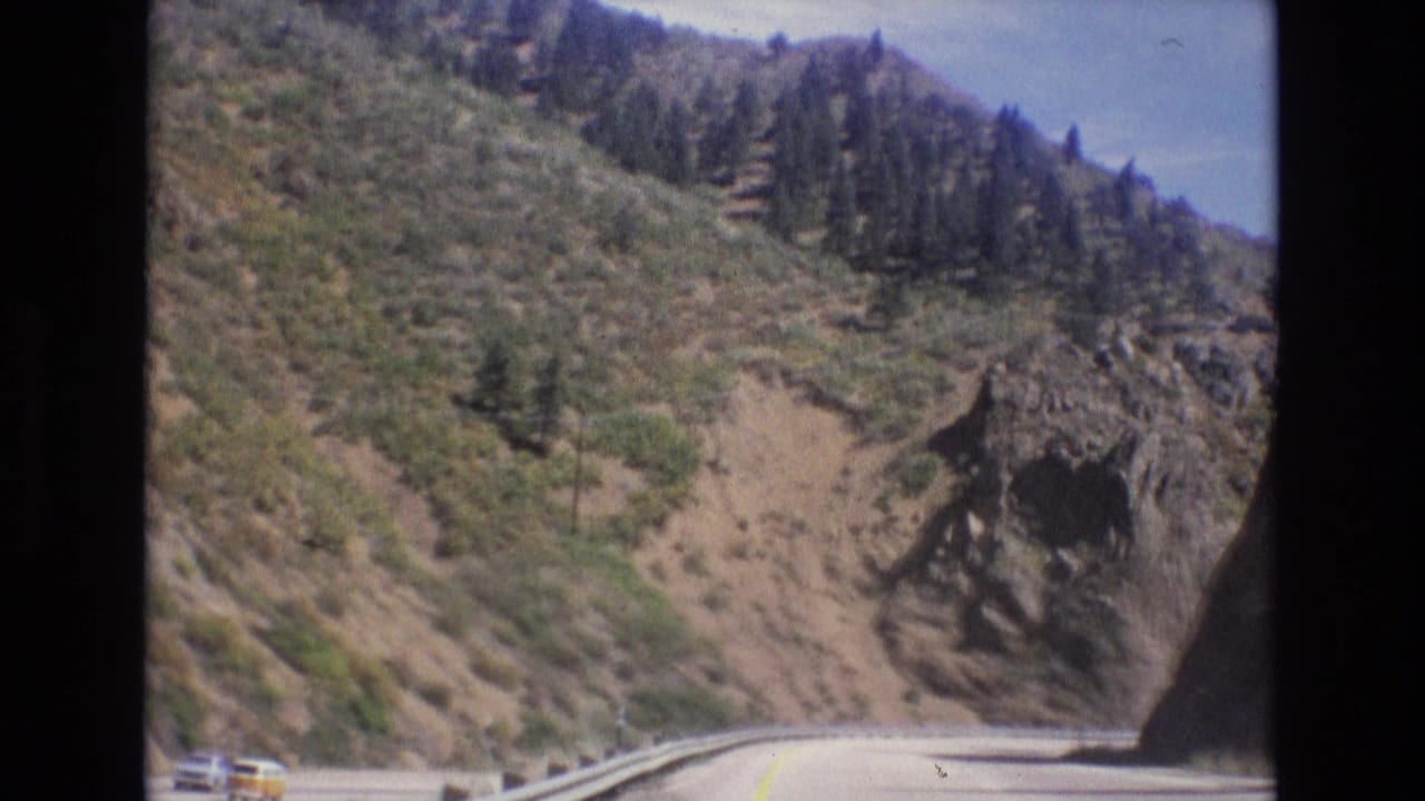 1973 Colorado 8mm frame from a moving vehicle showing a curving mountain highway, metal guardrail, rocky hillside, shrubs, pine trees, and a small yellow vehicle at the road edge.