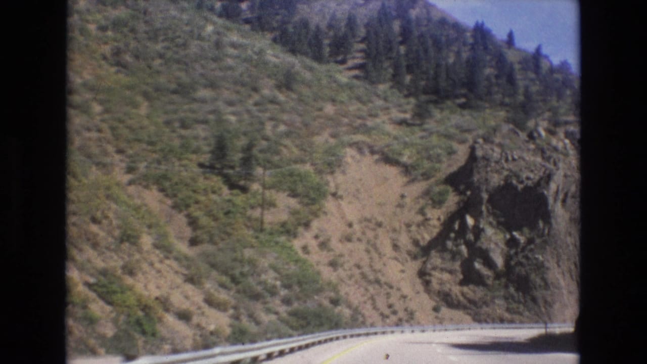 Mountain road curves beside a guardrail under a rocky hillside with trees.
