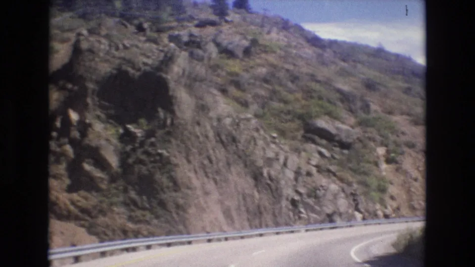 1973 Colorado 8mm frame with the moving camera passing a rocky roadside cut, sparse vegetation, metal guardrail, and the edge of a curving mountain highway.