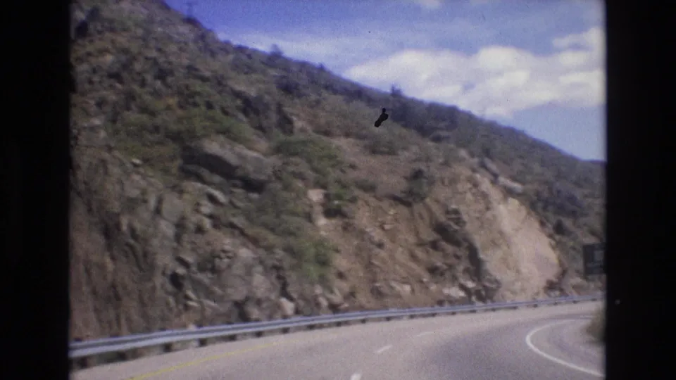 1973 Colorado 8mm frame looking ahead on a winding mountain road bordered by a metal guardrail, rocky slopes, sparse shrubs, distant hills, blue sky, and clouds.