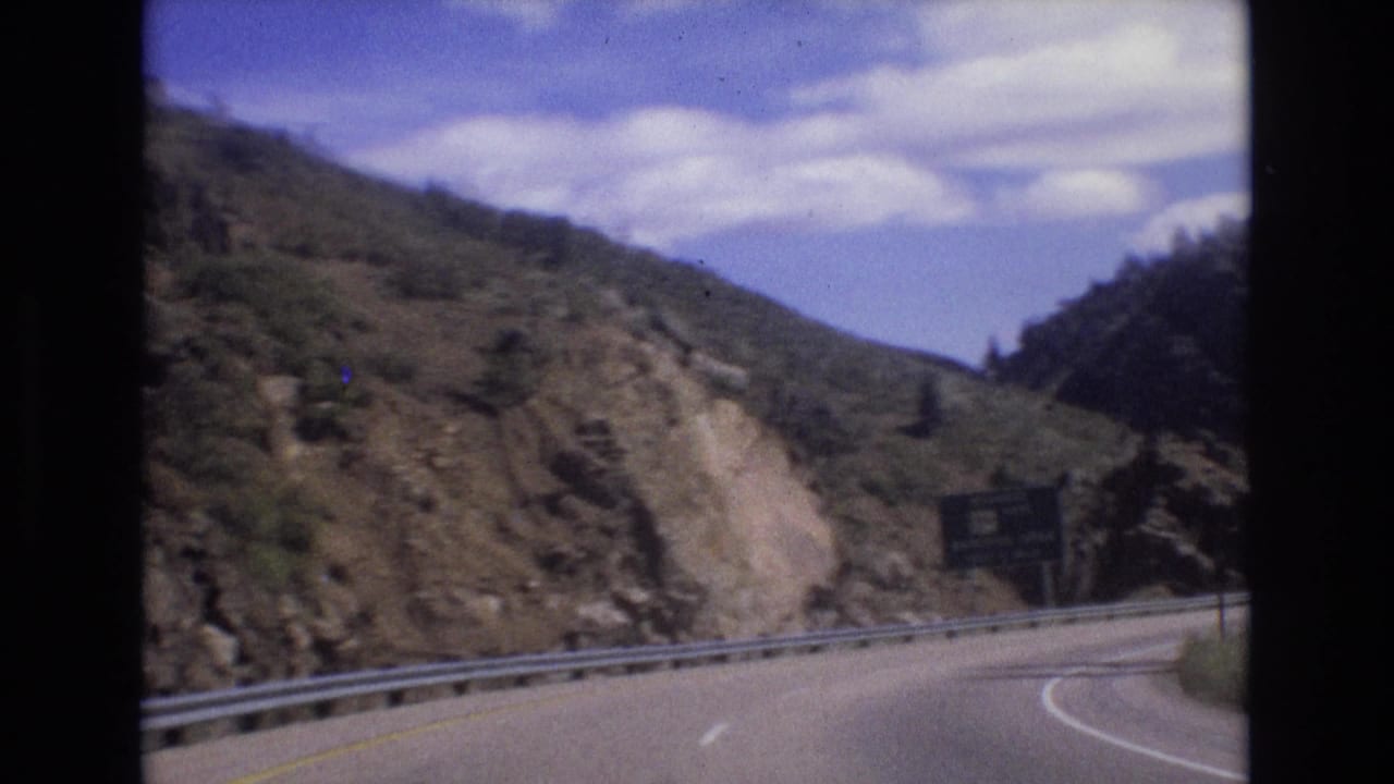 Curving mountain road with guardrail, rocky hillside, and a highway sign ahead.