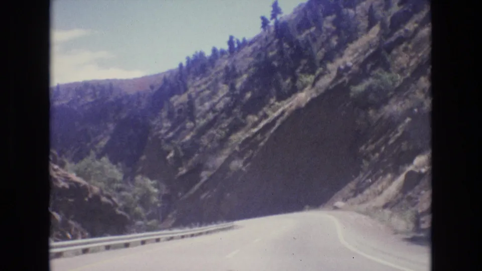 1973 Colorado 8mm frame looking along a bright curving mountain road with guardrail, rocky cut slope, sparse brush, pine trees, and distant canyon terrain.