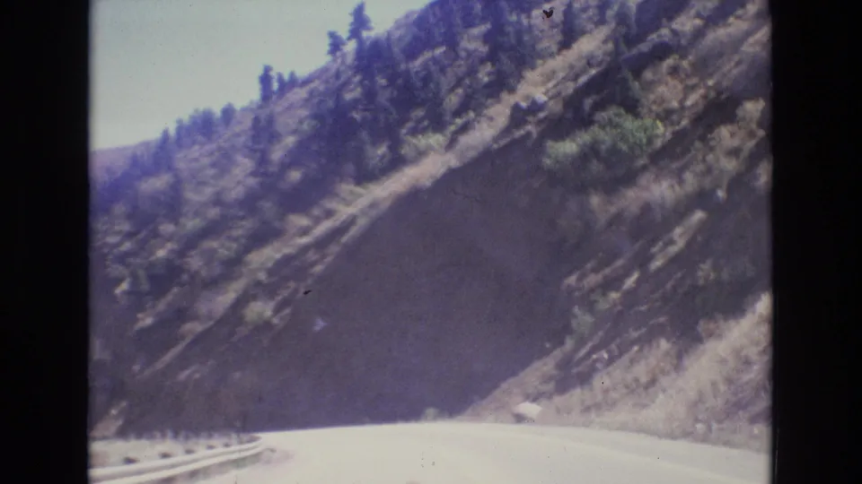 1973 Colorado home-movie frame from a moving vehicle, showing a sunlit mountain road, roadside guardrail, exposed rock slope, sparse vegetation, and pine trees along the canyon.
