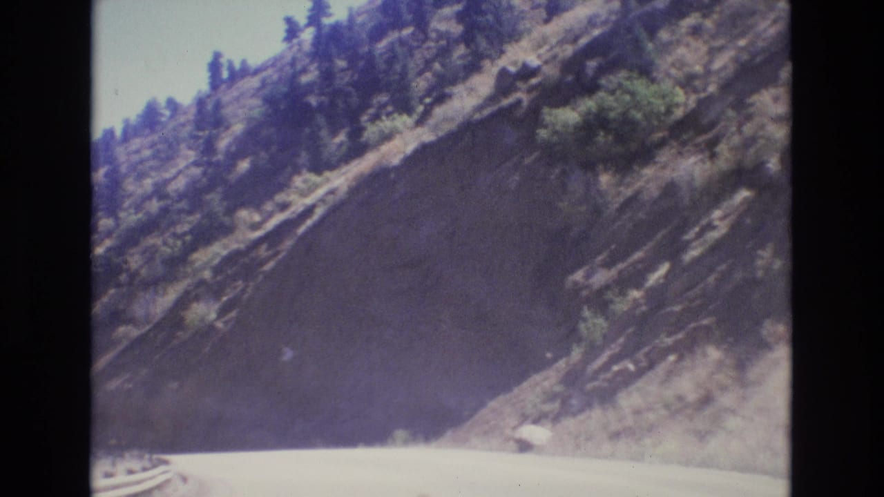 1973 Colorado 8mm home-movie frame showing a pale road curving along a steep rocky mountainside with scattered evergreens and bright sky.