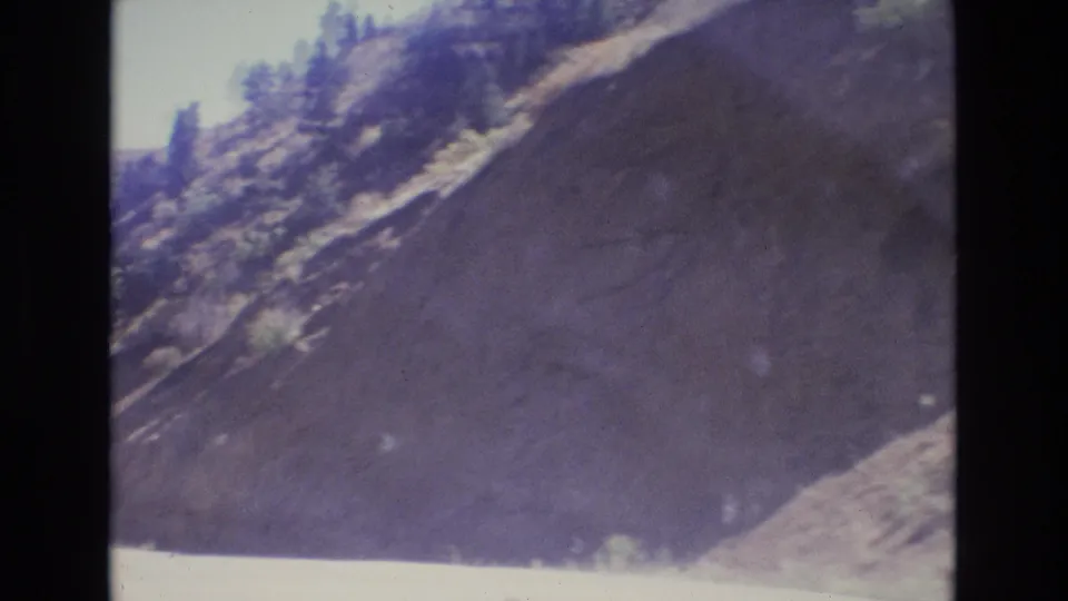 1973 Colorado travel footage frame with a moving-vehicle view of a bright road at the base of a steep canyon wall and sparse evergreen trees near the top.