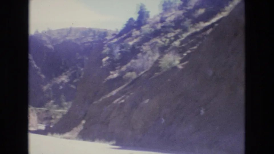 1973 Colorado 8mm frame showing a winding road through rocky mountain slopes, with a darker canyon backdrop and scattered trees softened by motion blur.