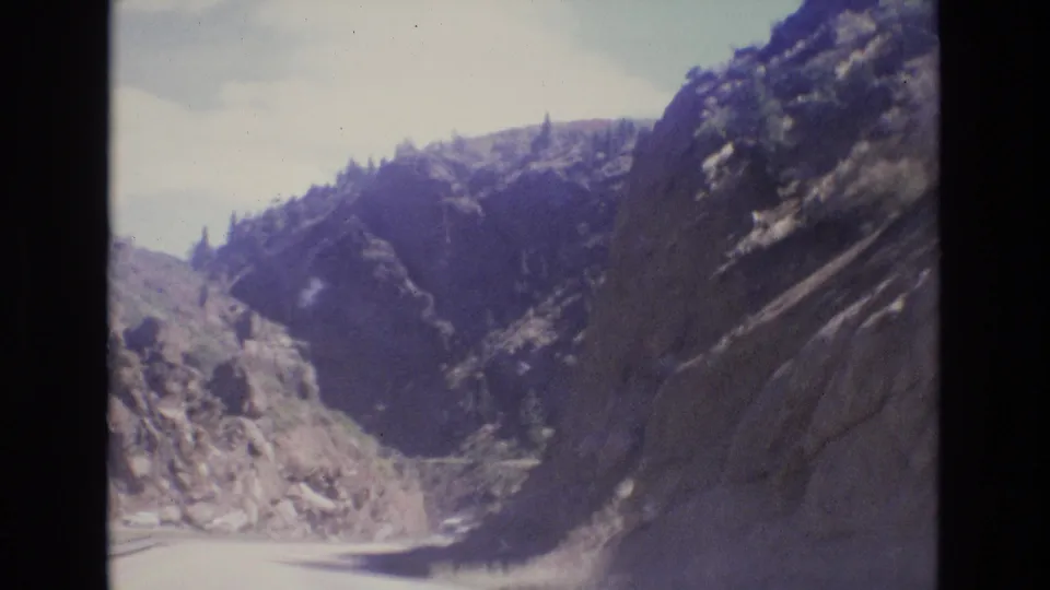 1973 Colorado home-movie frame looking ahead from a moving vehicle as the road enters a narrow canyon between rocky walls and forested upper slopes.