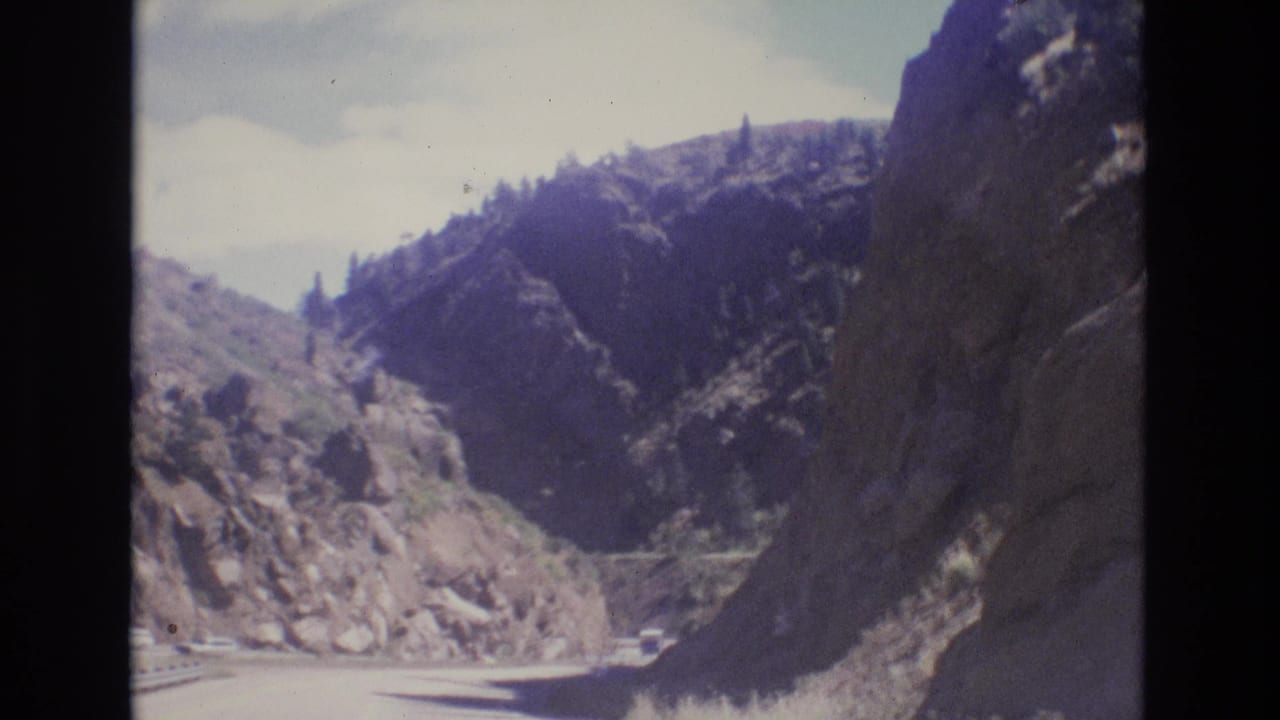 1973 Colorado 8mm frame showing a winding mountain road bordered by steep rocky canyon walls and sparse vegetation beneath a pale blue sky.