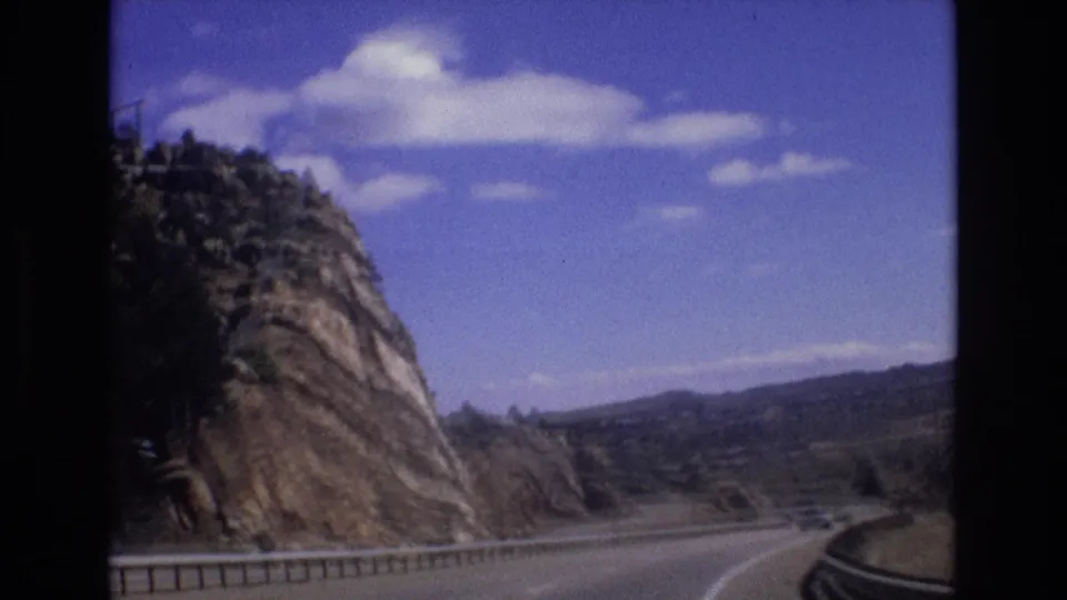 1973 Colorado 8mm frame from a moving vehicle, looking past a rocky cliff, guardrail, and curving road toward distant hills under scattered clouds.