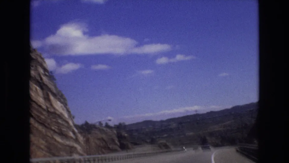1973 Colorado 8mm frame showing the road continuing past a rocky slope and guardrail toward distant hills, with a small vehicle barely visible ahead.