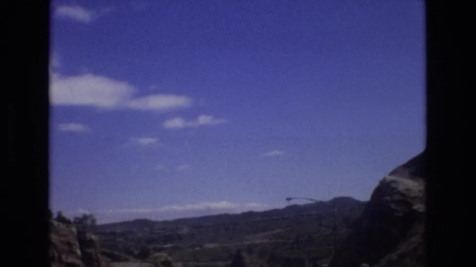 1973 Colorado 8mm frame looking across a mountain roadway toward distant hills, roadside poles, scattered clouds, and a rocky outcrop at the frame edge.