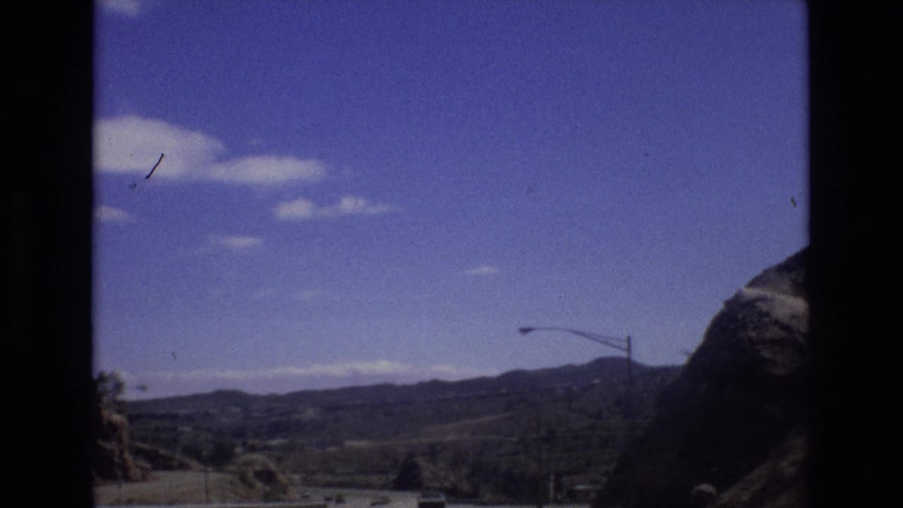 Highway viewed from a moving vehicle with mountains, guardrails, light poles, cars, and blue sky.