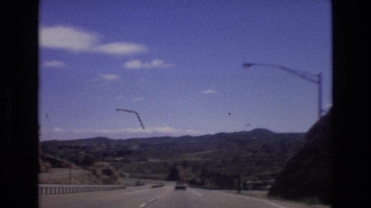 Roadway curves through a valley with cars ahead, guardrail, light poles, hills, and mountains.