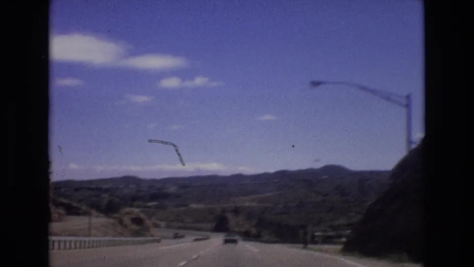 1973 Colorado road-view frame from a moving vehicle, looking down a highway with cars ahead, guardrail, streetlights, open valley land, and dark mountain ridges beyond.