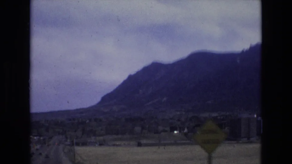 1973 Colorado 8mm frame showing a mountain range beyond low roadside development, open ground, a road edge, and a blurred sign seen from a moving vehicle.