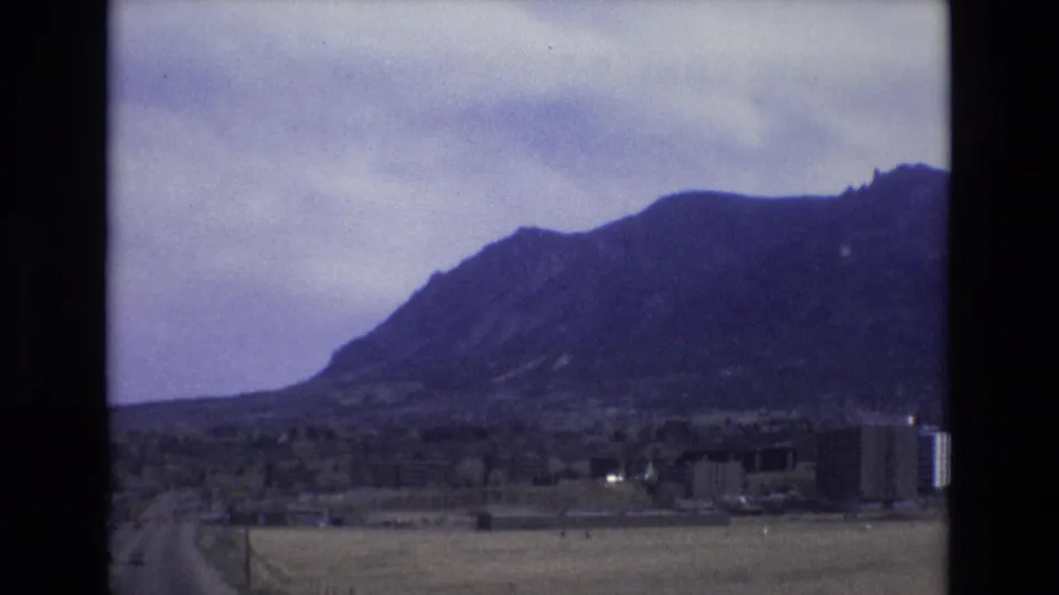 1973 Colorado home-movie frame showing a broad mountain slope behind open roadside land, small buildings, and a roadway edge under a pale cloudy sky.