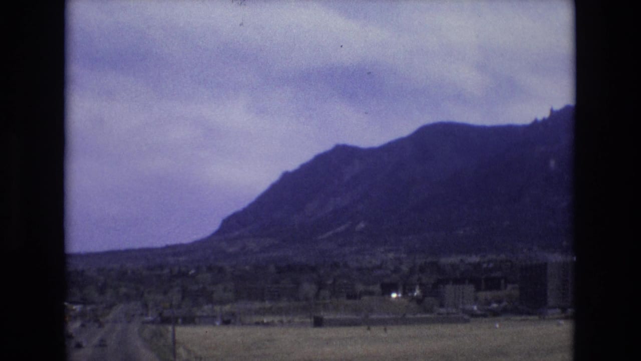 1973 Colorado 8mm frame showing a rugged mountain range beyond open fields, a roadside foreground, utility poles, and scattered low buildings under a cloudy sky.