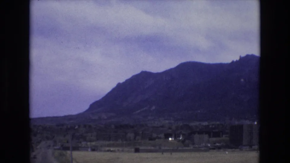 1973 Colorado home-movie frame with a dark mountain slope, cloudy sky, open roadside land, and a few distant buildings seen from a moving vehicle.