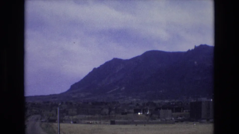 1973 Colorado 8mm frame showing open fields and roadside poles in front of a broad mountain range, with distant buildings softened by home-movie focus and grain.