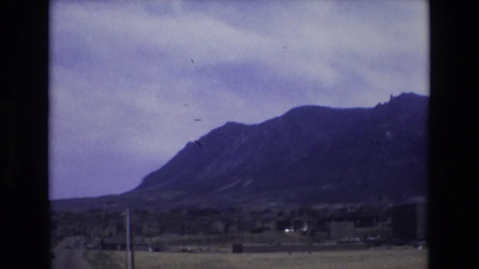 1973 Colorado travel footage frame showing a cloudy mountain landscape, sparse roadside poles, open ground, and distant low buildings at the edge of the frame.