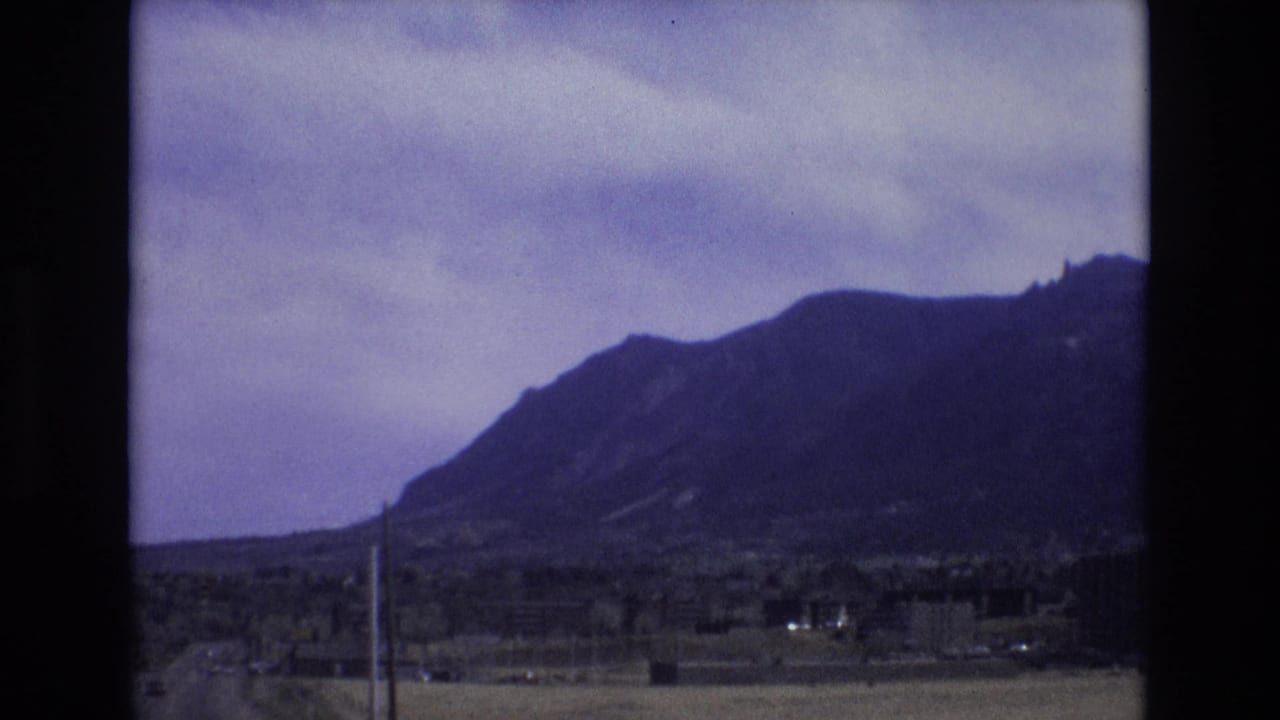 1973 Colorado 8mm frame showing a dark mountain ridge beneath cloudy sky, with sparse roadside or suburban foreground and utility poles.