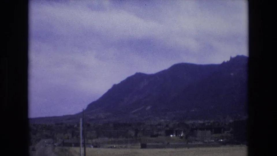 1973 Colorado 8mm frame showing a dark mountain ridge beneath cloudy sky, with sparse roadside or suburban foreground and utility poles.