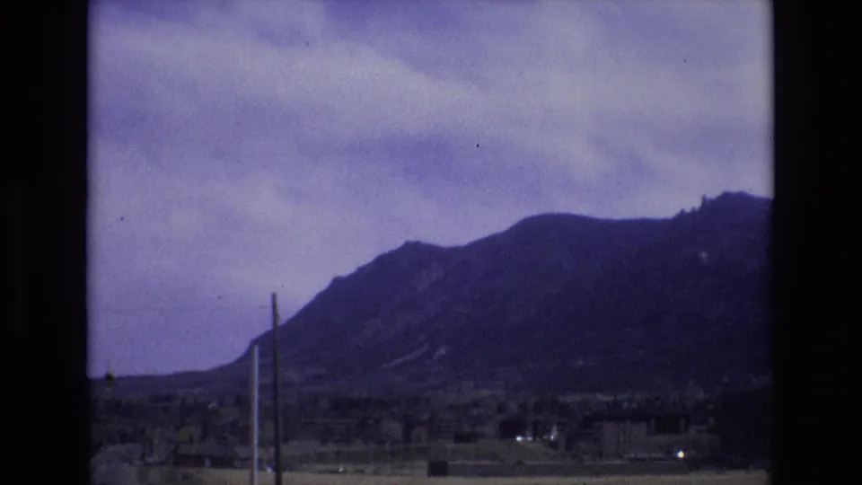 1973 Colorado home-movie frame with a mountain range, muted cloudy sky, low structures, and utility poles crossing the foreground.