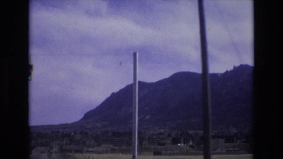 1973 Colorado home-movie frame with a dark mountain slope, overcast sky, low foreground structures, and prominent vertical poles near the lens.