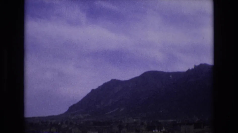 1973 Colorado 8mm frame with a prominent mountain slope, low roadside foreground, cloudy sky, and visible dark film-frame edge.