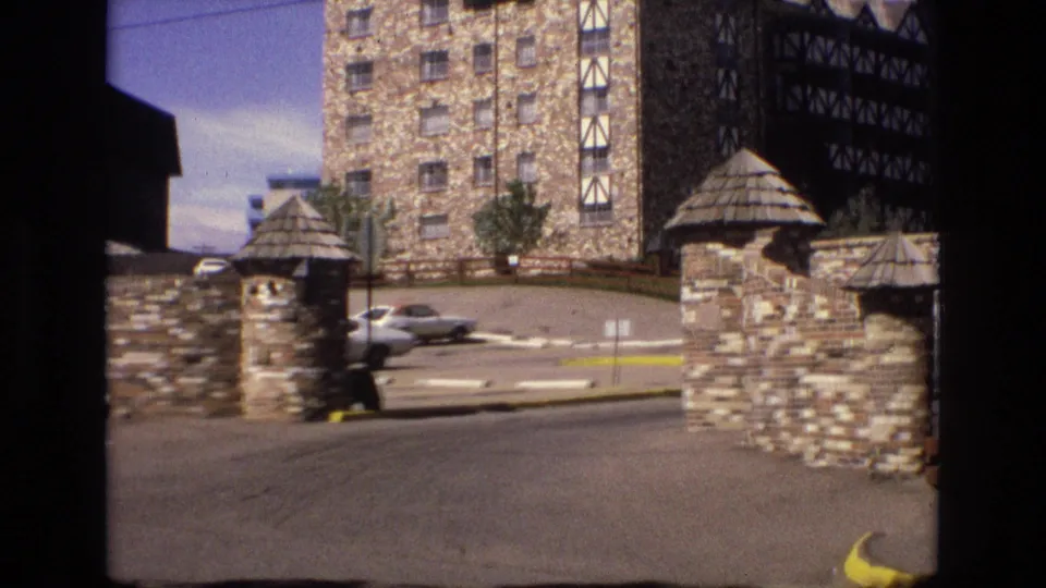 1973 Colorado 8mm frame showing a stone-and-timber building entrance with masonry gate posts, parked cars, paved drive, and mountain-sky background glimpses.