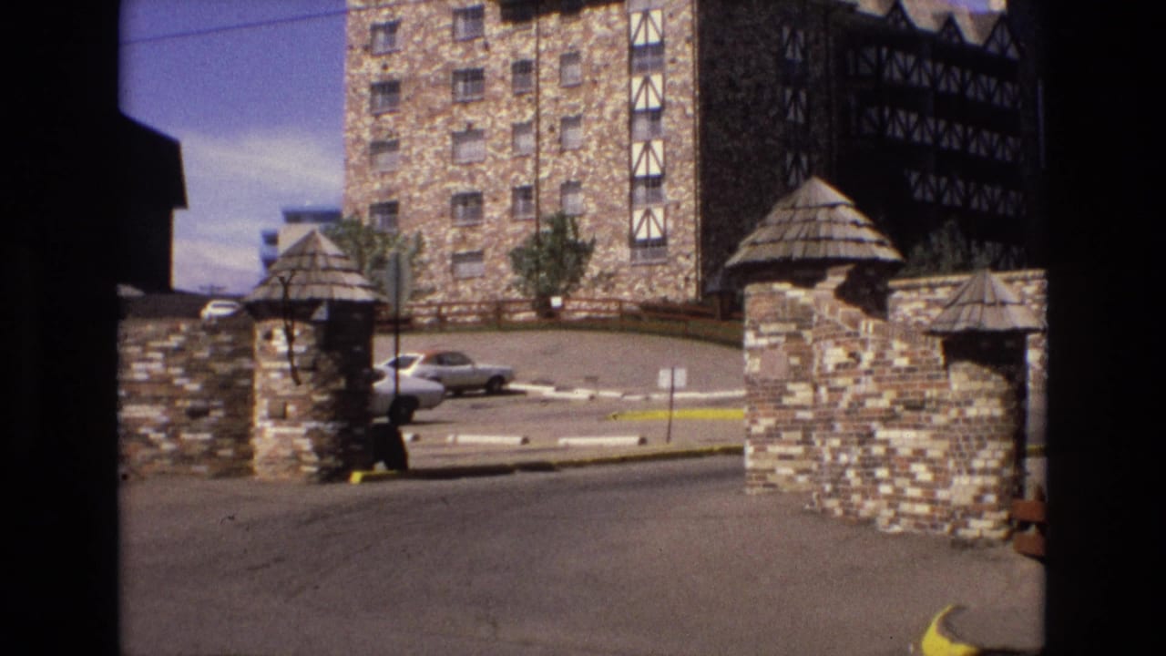 1973 Colorado 8mm frame showing a stone-and-timber lodge entrance with conical-roof gate structures, a paved driveway, yellow curb, and a parked sedan in the background.