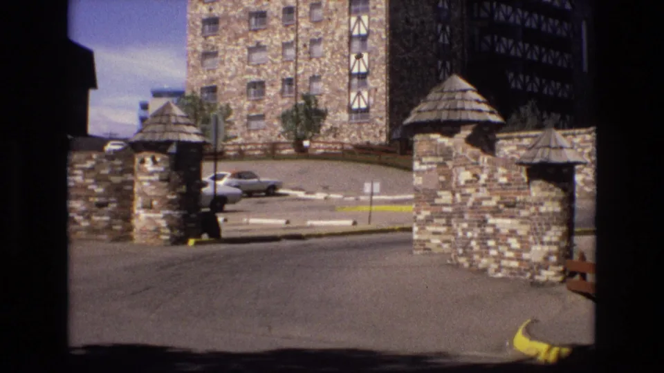 1973 Colorado home-movie frame of a stone lodge entrance, showing the gate structure, driveway, yellow curb markings, parked cars, and textured stone-and-timber facade.