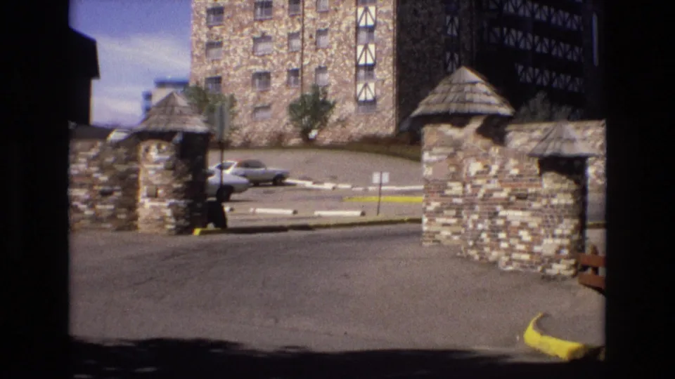 1973 Colorado 8mm frame with a closer visitor view of the stone entrance gate, paved drive, yellow curb, parked sedan, and lodge facade beyond.