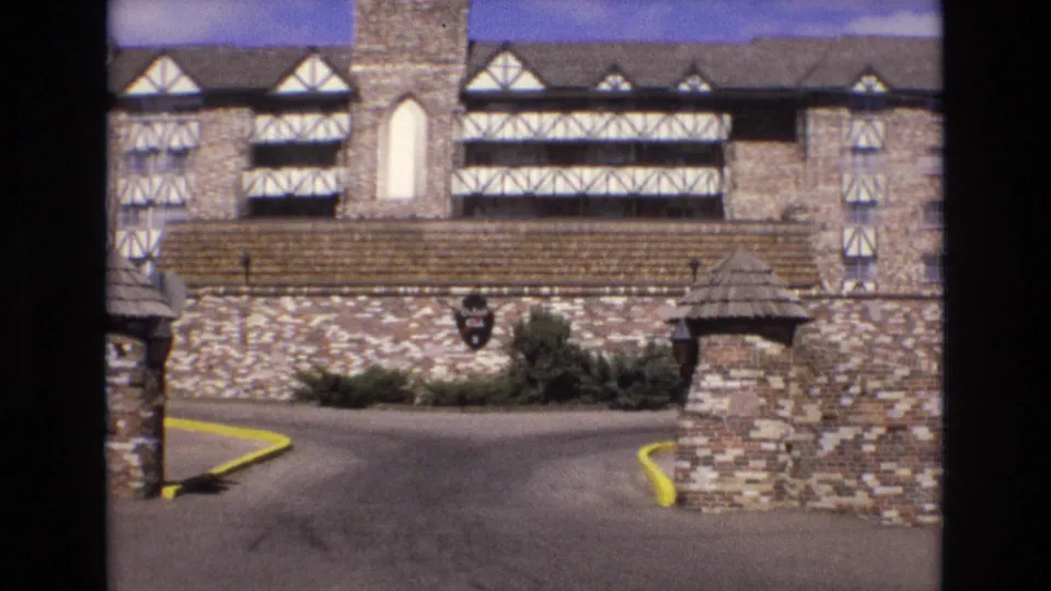 1973 Colorado 8mm frame showing the lodge facade more directly, with half-timbered upper walls, stonework, entrance gate walls, shrubs, a paved drive, and yellow curbs.