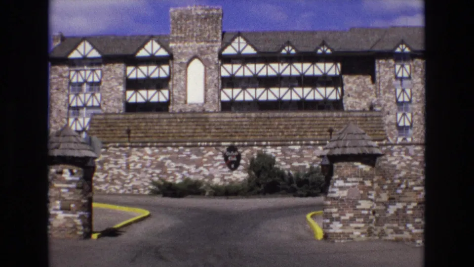 1973 Colorado 8mm view of a Tudor-style hotel or club exterior with stonework, decorative timber framing, a walled driveway entrance, shrubs, and yellow-painted curbs.