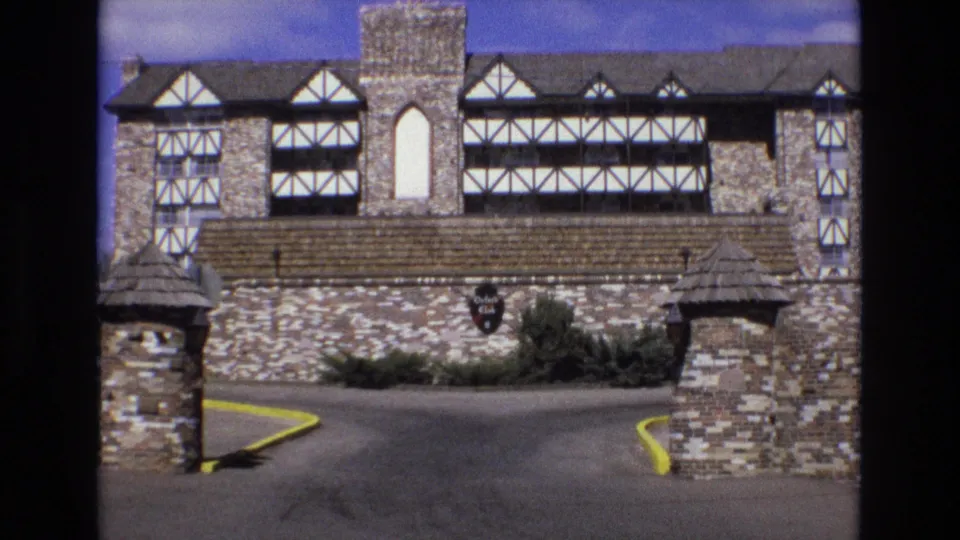 1973 Colorado frame showing a steady exterior view of a Tudor-style stone hotel or club property, including brick gateposts, a paved private drive, yellow curbs, shrubs, and a mounted sign.