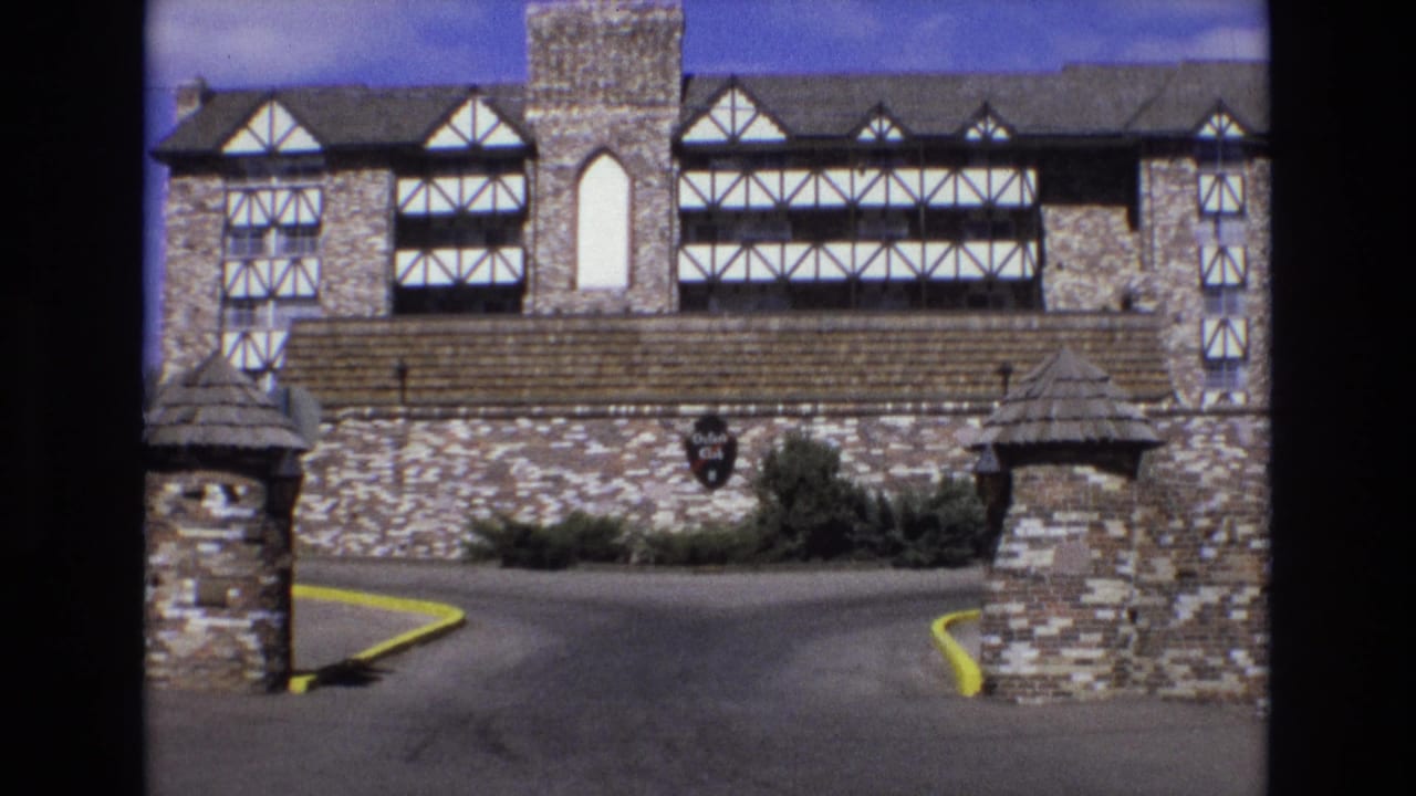 1973 Colorado 8mm frame showing a Tudor-influenced stone building beyond a driveway entrance with stone piers, yellow curbs, and a shield sign on the wall.