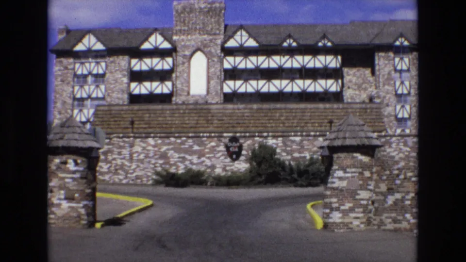 1973 Colorado 8mm frame showing a Tudor-influenced stone building beyond a driveway entrance with stone piers, yellow curbs, and a shield sign on the wall.