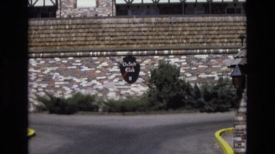 Closer 1973 Colorado 8mm frame showing the Oxford Club shield sign on a stone wall beneath a shingled roof edge, with shrubs and the entrance drive in front.