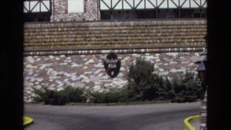 Final 1973 Colorado frame from the approach, centered on the stone wall and readable Oxford Club sign with shrubbery, driveway pavement, and half-timbered facade above.