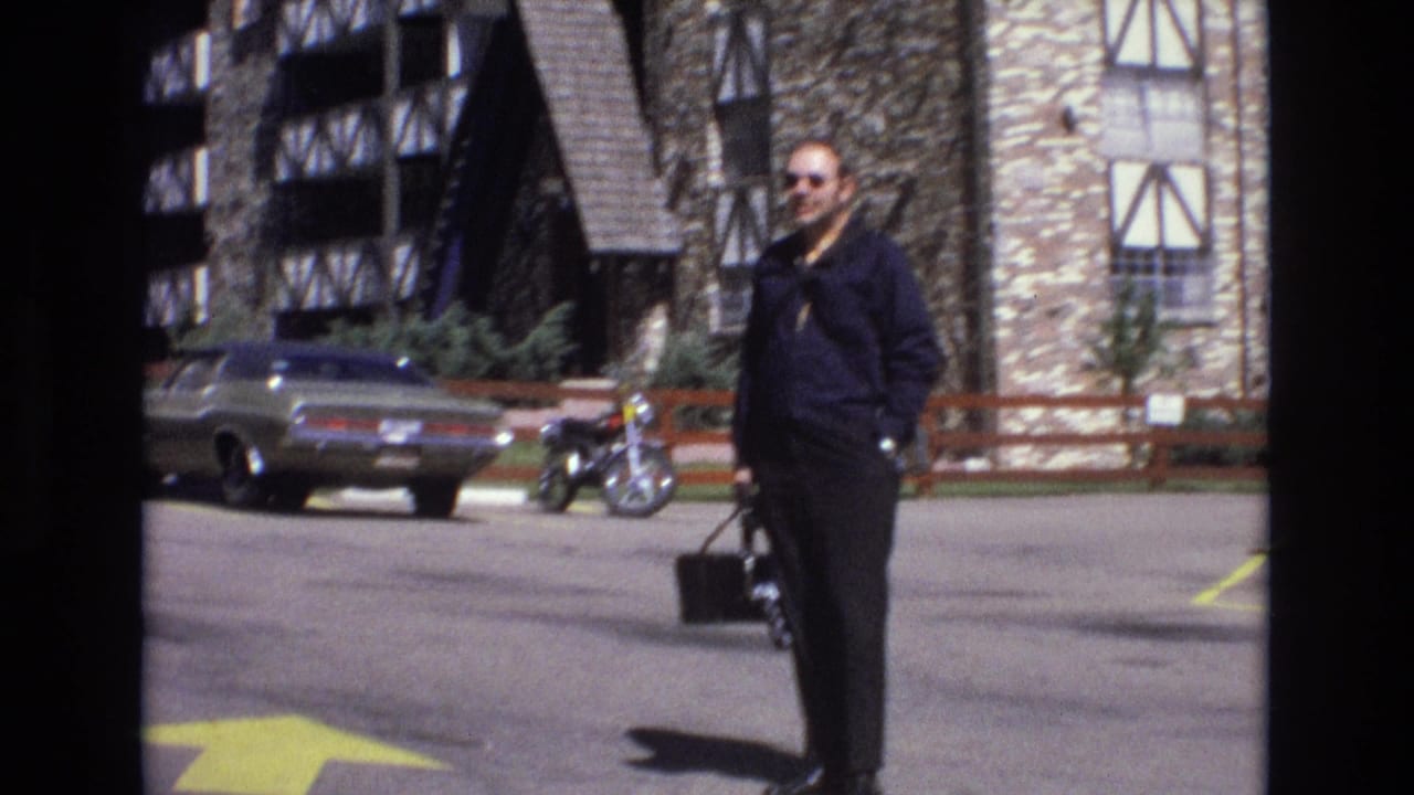 Man in sunglasses stands in a parking lot holding a black bag near a parked car and motorcycle.